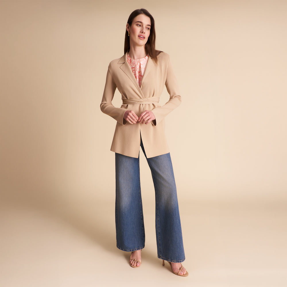 Woman wearing a beige blazer over a light blue shirt and jeans on a plain background