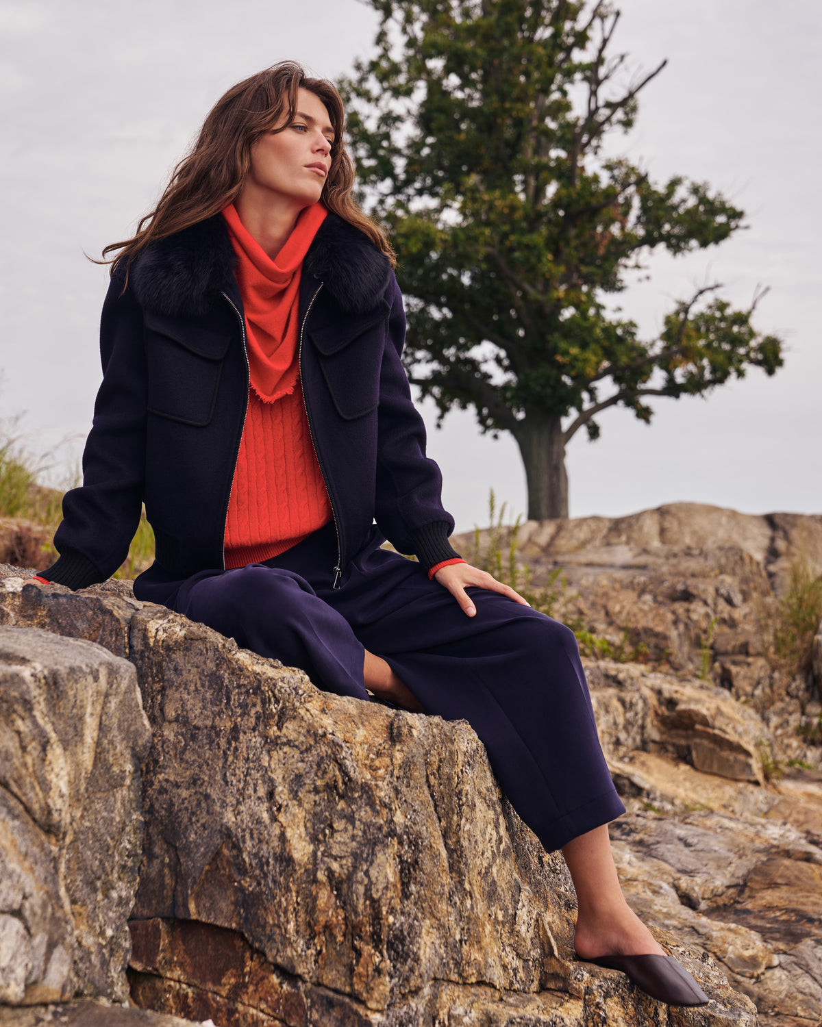 Woman in a navy coat and rust sweater from the M.M.LaFleur October collection, seated on coastal rocks