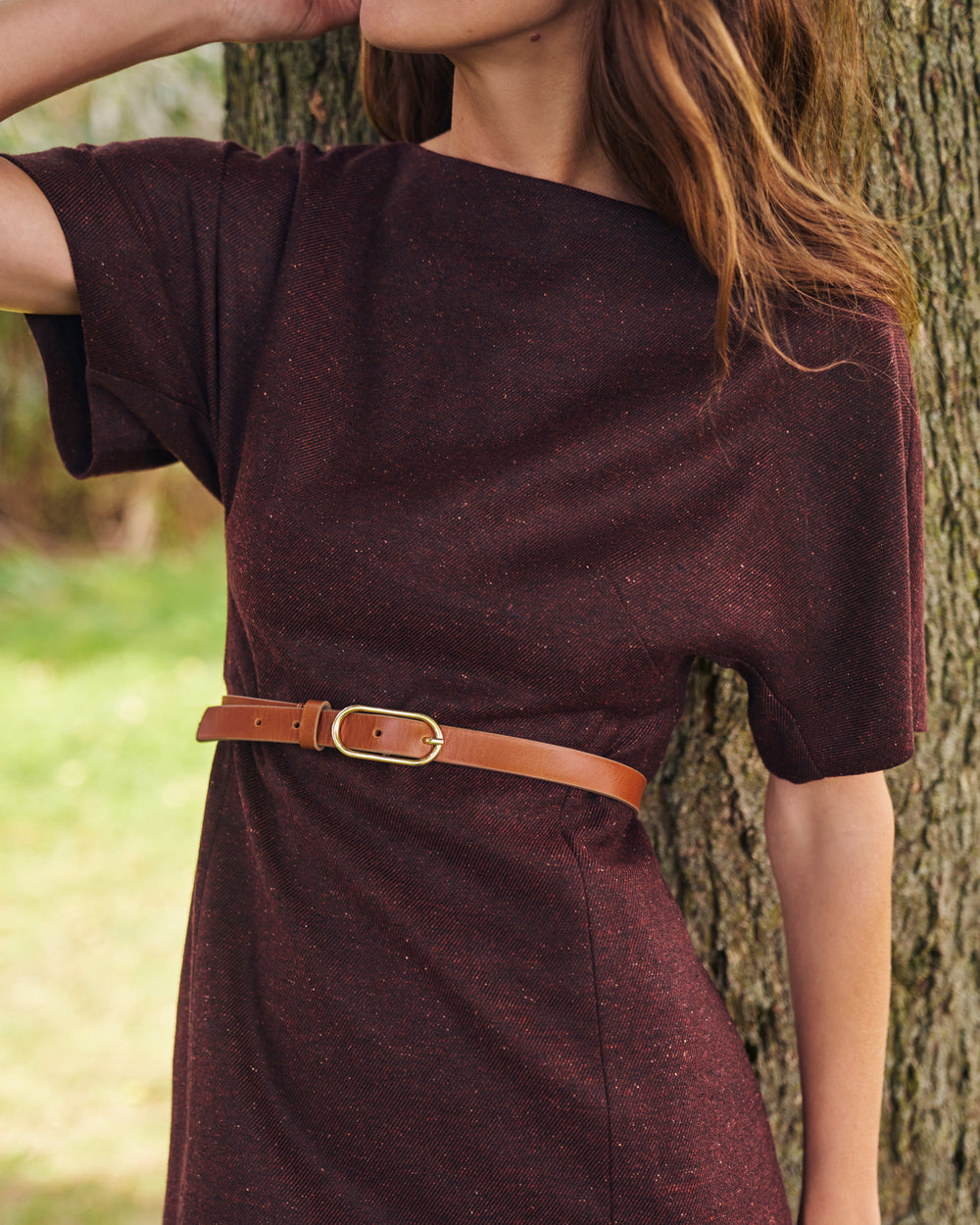 A woman wearing a short-sleeve dark burgundy dress with a thin brown leather belt, standing outdoors next to a tree trunk.