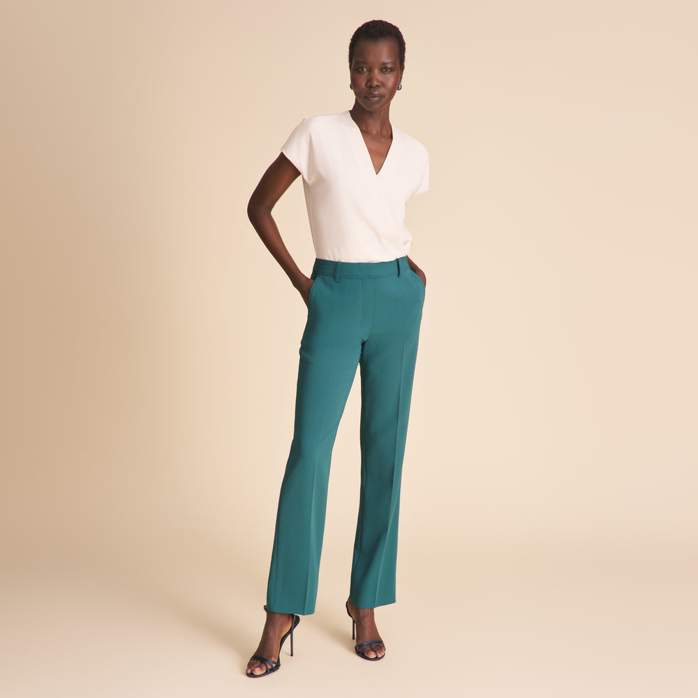 Woman wearing a white top and teal pants against a beige background
