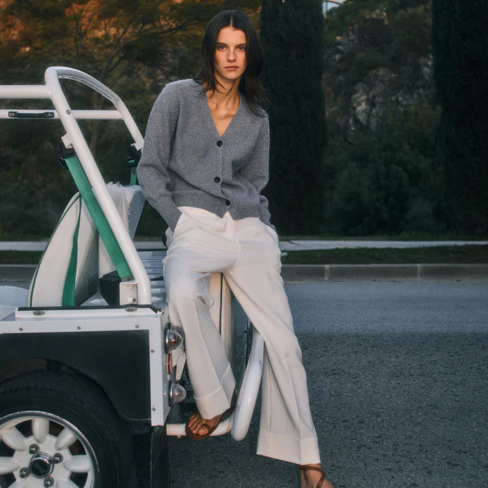 Woman in gray cardigan and white pants leaning against a vehicle with a scenic background