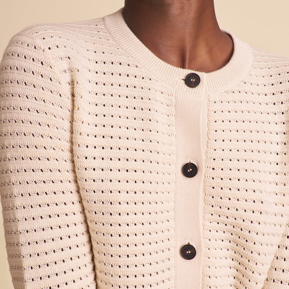 Close-up of a person wearing a beige knitted cardigan with black buttons on a beige background